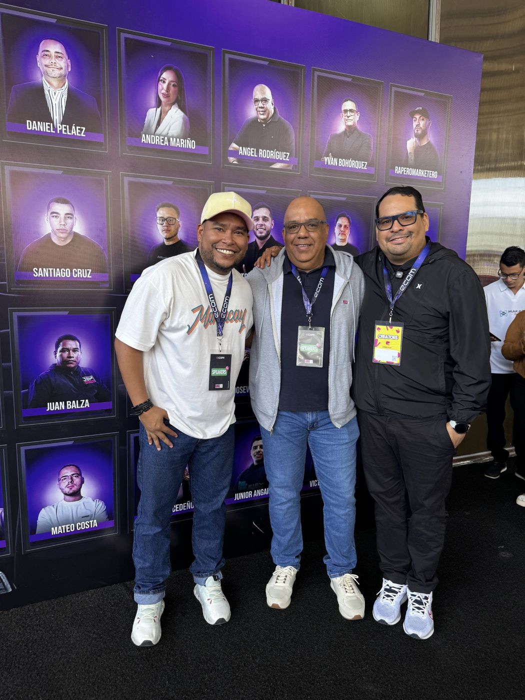 Juan Balza con dos colegas frente al muro de speakers de eCom Venezuela, donde aparece su foto como ponente